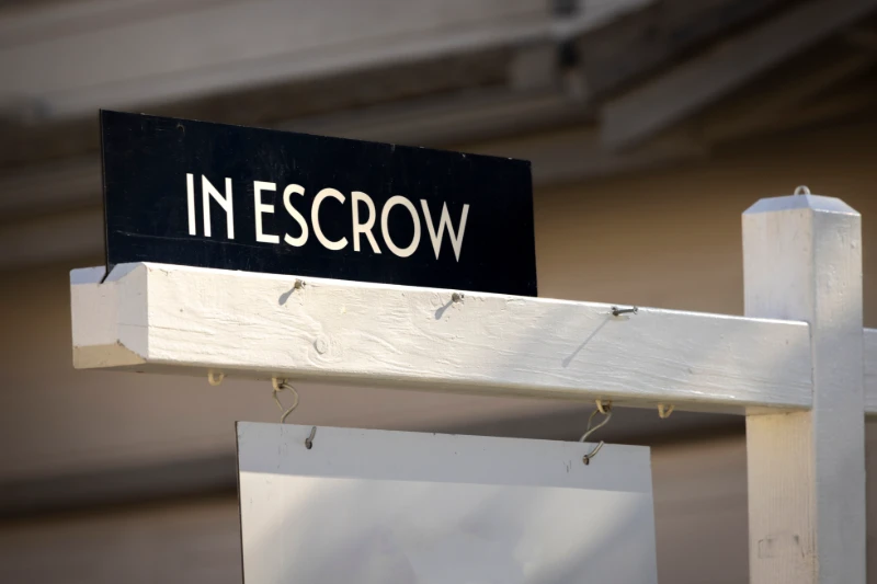 A close-up of a real estate signpost with a black sign reading “In Escrow,” indicating a pending property sale; the background is out of focus.