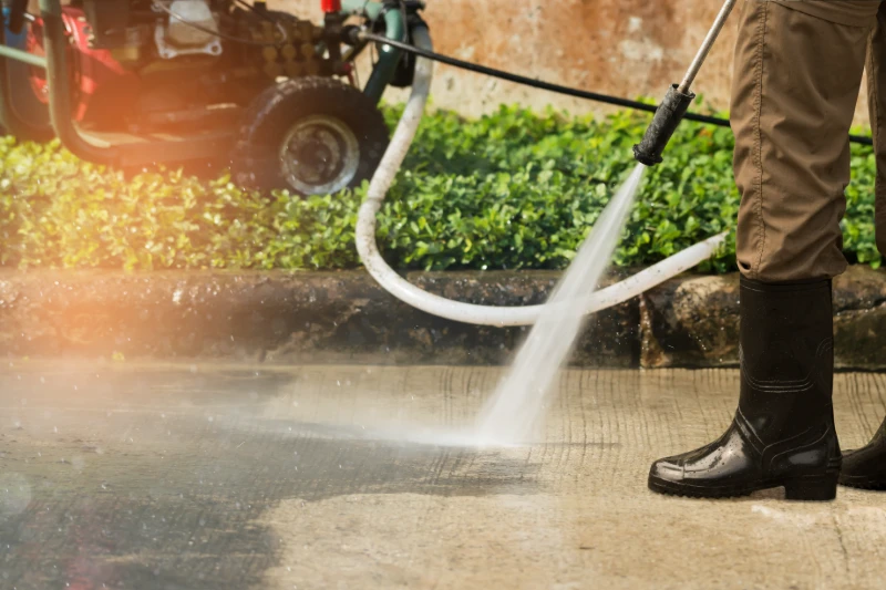 A person in rubber boots uses a pressure washer to clean a concrete surface outdoors, with green plants and equipment visible in the background.