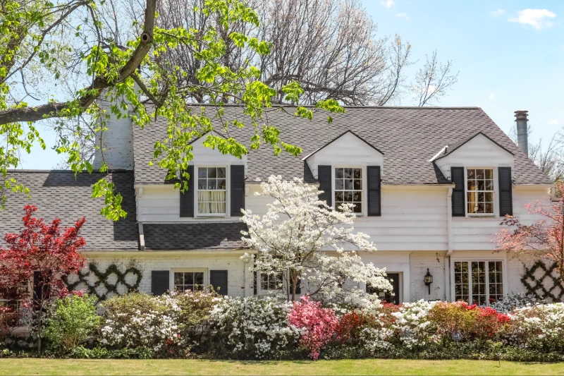 A white two-story house with black shutters is surrounded by blooming trees and colorful bushes in a lush, green yard, under a clear blue sky.