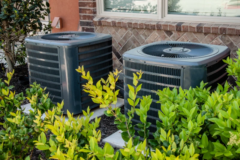 Two outdoor air conditioning units sit beside a brick house, partially surrounded by green bushes and plants, with a window visible above the units.