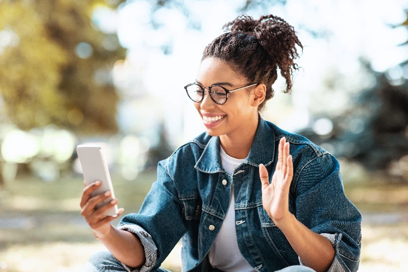 A young woman wearing glasses and a denim jacket sits outdoors, smiling and waving at her smartphone during a video call. Trees and sunlight are visible in the blurred background.