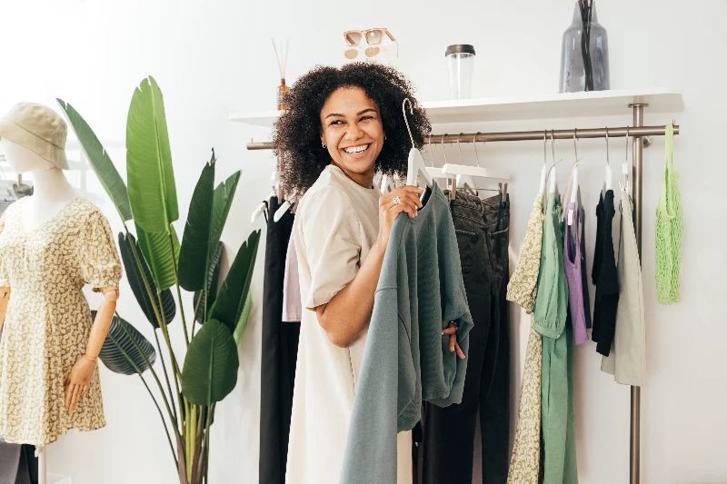 A smiling woman holds a green sweatshirt while standing in a modern clothing store, surrounded by hanging clothes, plants, and a mannequin in a floral dress.