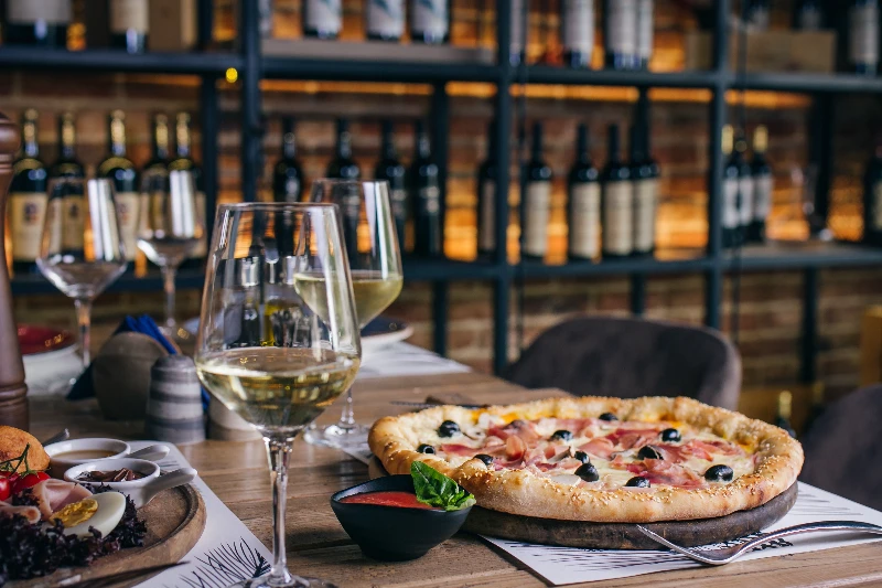 A table set in a restaurant with a thin-crust pizza, two glasses of white wine, a small bowl of sauce, and appetizers. Shelves with numerous wine bottles line the brick wall in the background.