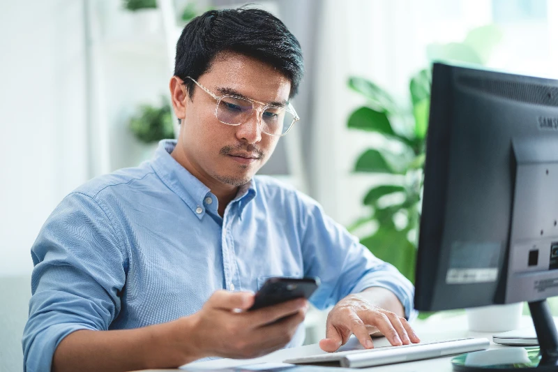 A man wearing glasses and a light blue shirt sits at a desk, looking at his smartphone while using a desktop computer. There are green plants in the background.