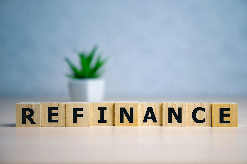 Wooden blocks spelling REFINANCE are arranged in a row on a table, with a small potted plant blurred in the background.