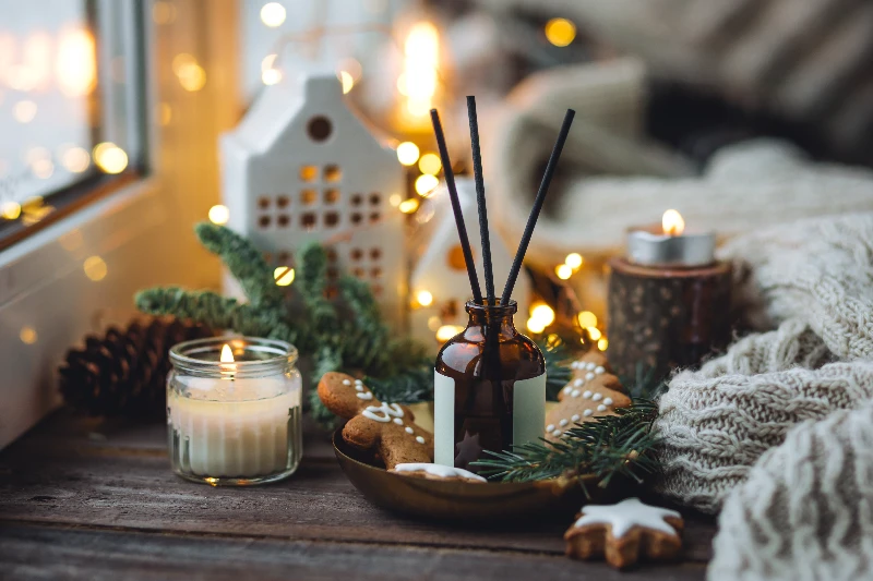 A cozy winter scene with candles, gingerbread cookies, pine branches, a knitted blanket, and a house-shaped lantern on a wooden surface, decorated with warm fairy lights.