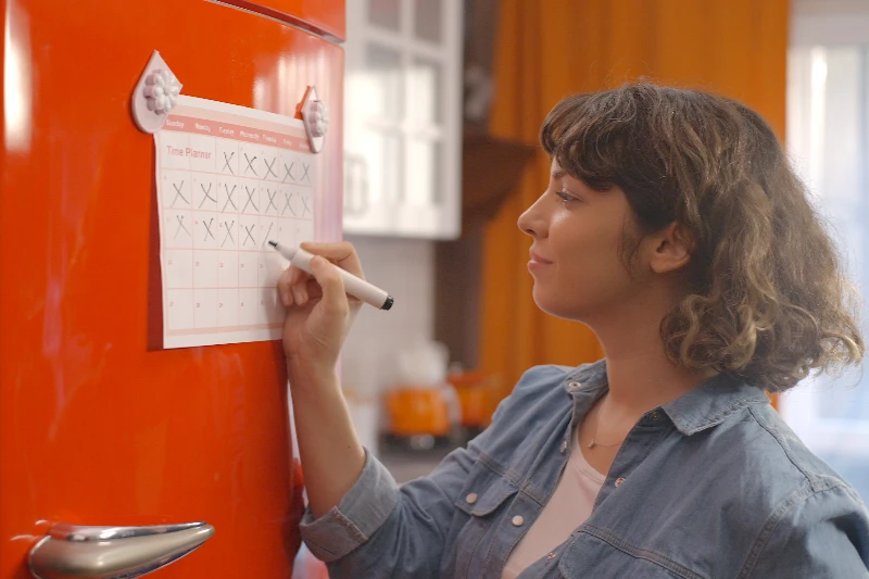 A woman marks a date on a calendar attached to a bright red refrigerator in a kitchen, using a black marker. She appears focused, and the kitchen has an orange and white color scheme.