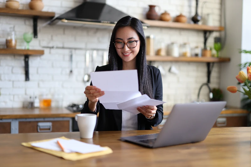 A woman wearing glasses and a blazer sits at a kitchen table, smiling as she reads a letter. A laptop, coffee mug, and documents are on the table, with shelves and kitchen items in the background.