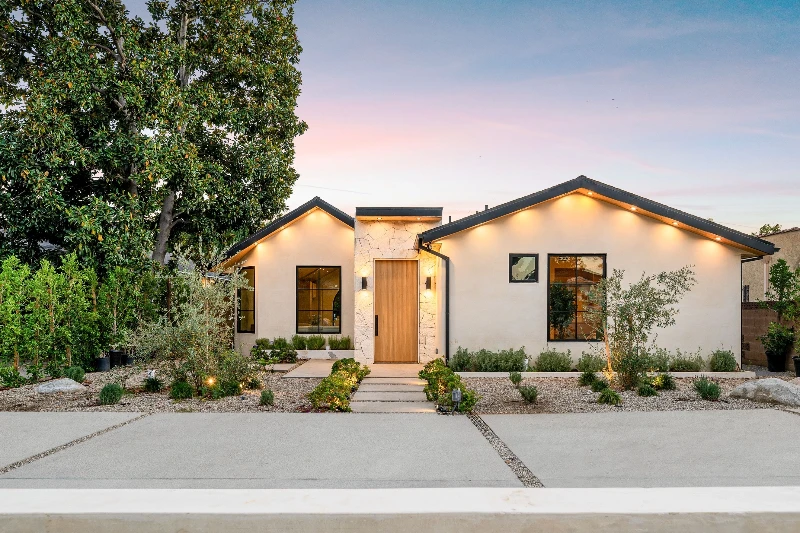 Modern single-story house with a flat front yard, light-colored exterior, large windows, wooden front door, pathway, landscaped plants, and exterior lights on at dusk. A tall tree stands to the left of the house.