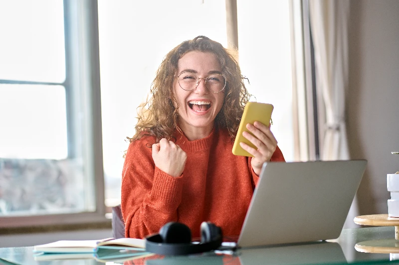 A smiling woman wearing glasses and an orange sweater sits at a desk with a laptop, holding a yellow phone and raising her fist in excitement. Light streams in through a window behind her.