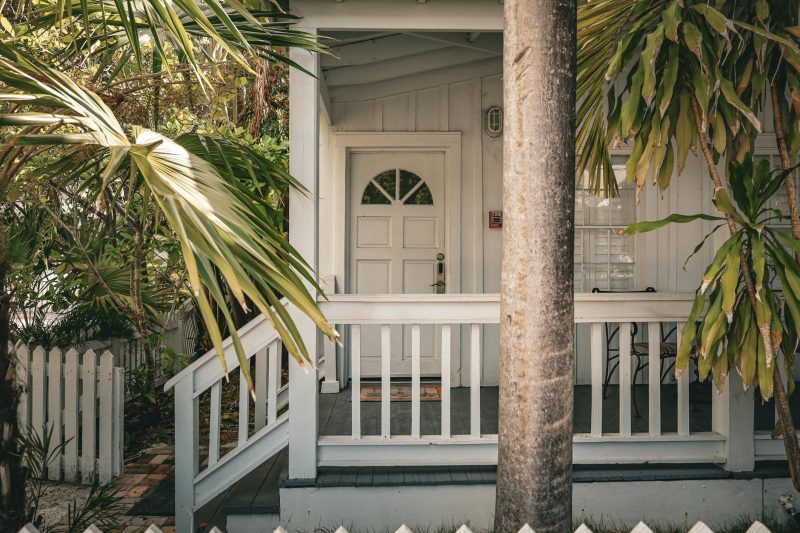 A white wooden house with a porch and front door is partially shaded by palm trees and surrounded by lush greenery, with a white picket fence in the foreground.