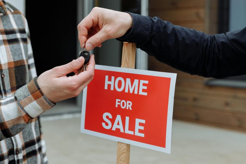Close-up of two people exchanging house keys in front of a red Home For Sale sign, indicating the sale or purchase of a home. Only their hands and part of their arms are visible.