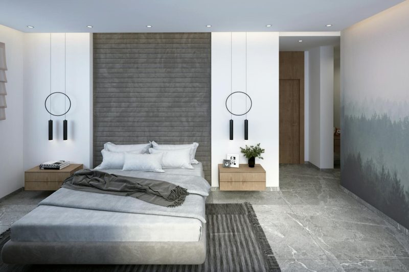 Modern bedroom with a gray upholstered bed, gray bedding, hanging pendant lights, wooden nightstands, and a textured accent wall. The floor is tiled, and a rug is placed beside the bed.