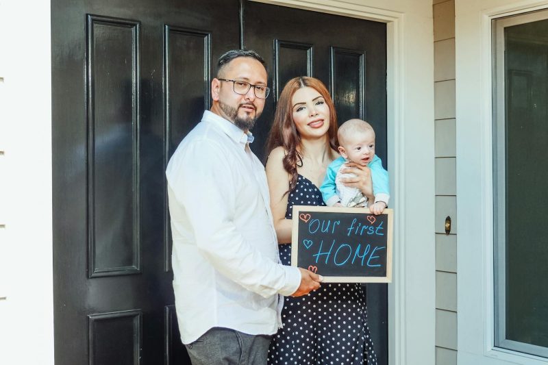 A man, woman, and baby stand in front of a black door. The woman holds a chalkboard sign that reads our first HOME with hearts. They are smiling, celebrating moving into their new home.