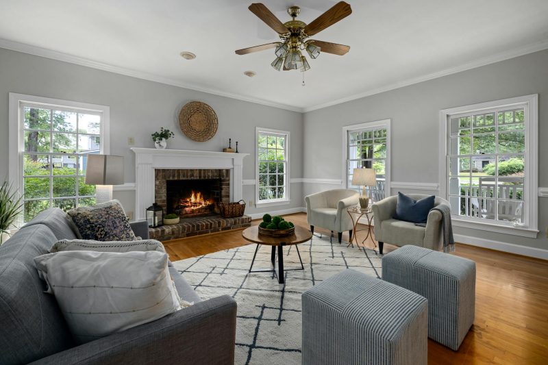 A bright living room with a gray sofa, armchairs, ottomans, a round coffee table, and a lit fireplace. Large windows let in natural light and offer views of greenery outside. The room has light gray walls and wooden floors.