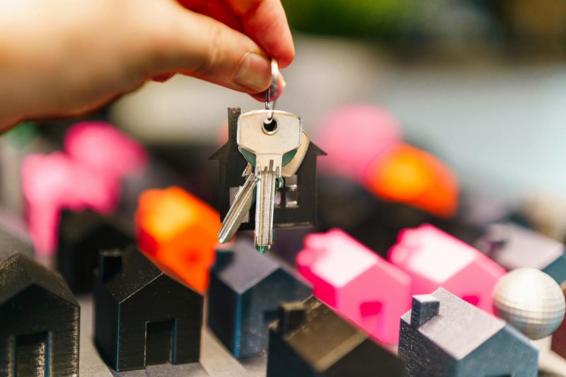 A hand holds a keyring with house-shaped keys above a surface with miniature model houses in black, pink, and orange colors, representing real estate or home ownership.