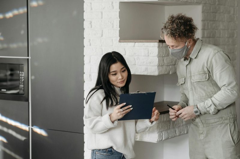 A woman holding a clipboard explains something to a man wearing a face mask, who is holding a pen. They are standing indoors near white brick shelves.