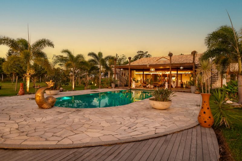 A serene backyard with a stone patio, oval swimming pool, tropical plants, decorative pots, and a wooden deck. A cozy, illuminated house with an outdoor seating area is in the background at sunset.