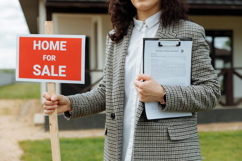 A woman holding a Home For Sale sign and a clipboard with documents stands in front of a house. She is wearing a checkered blazer and white shirt. The house is visible in the background.