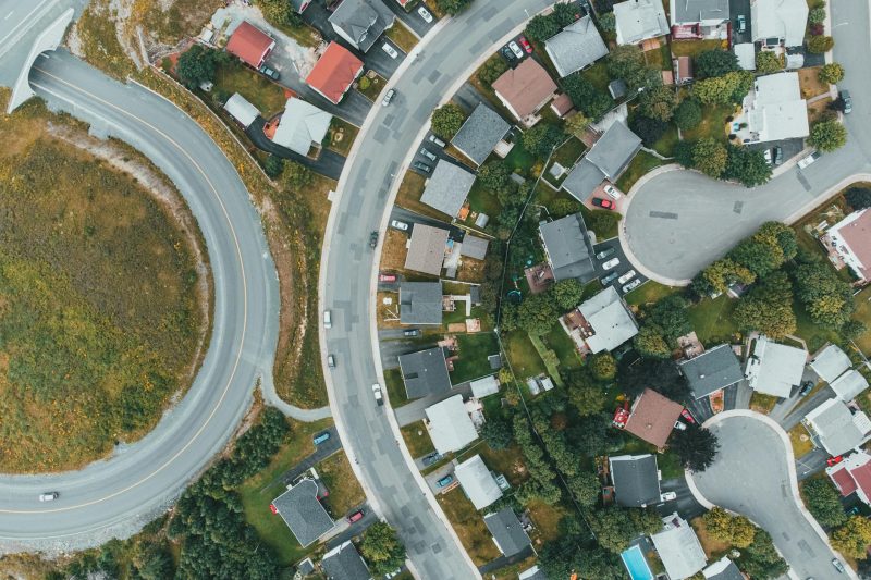 Aerial view of a suburban neighborhood with houses, lawns, and curving roads, including a large circular road on the left and residential streets lined with trees and parked cars on the right.