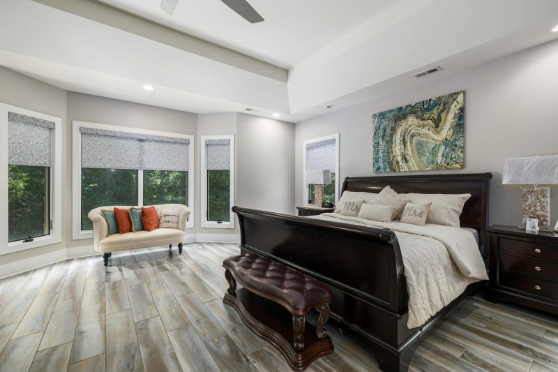 Spacious, modern bedroom with wood-look tile floors, a dark wood sleigh bed, a matching bench, and a beige loveseat beneath large windows with gray shades. Abstract artwork hangs above the bed.