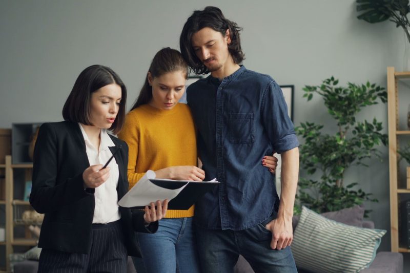 A professional woman shows documents to a couple in a living room. The couple stands close together, looking at the papers attentively. Houseplants and shelves are visible in the background.