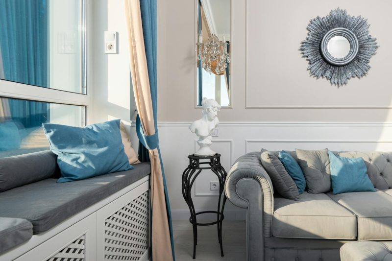 A bright, elegant living room with a gray couch, blue and gray pillows, a window seat, a classical bust on a black stand, a sunburst mirror, and a chandelier on the wall. Blue curtains frame large windows.