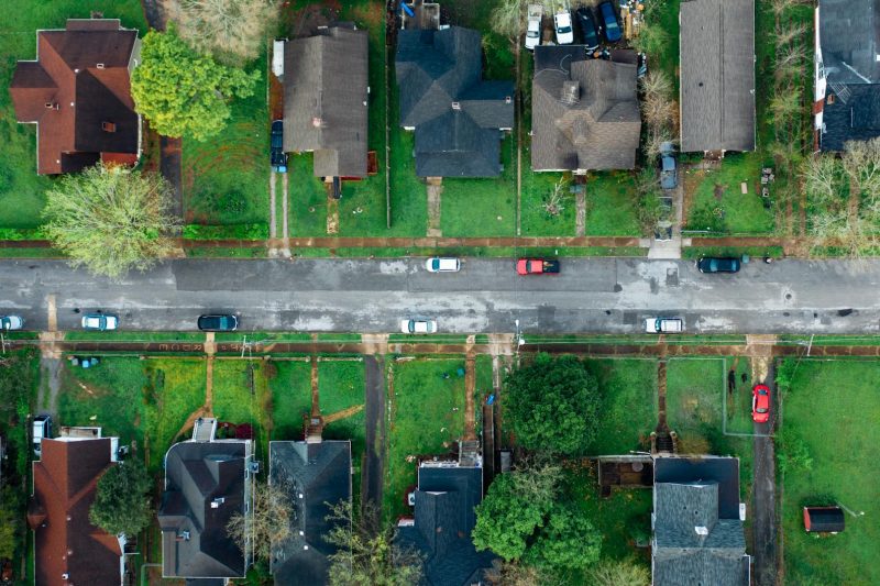 Aerial view of a suburban neighborhood showing two rows of houses separated by a street with several parked cars, green lawns, and scattered trees.