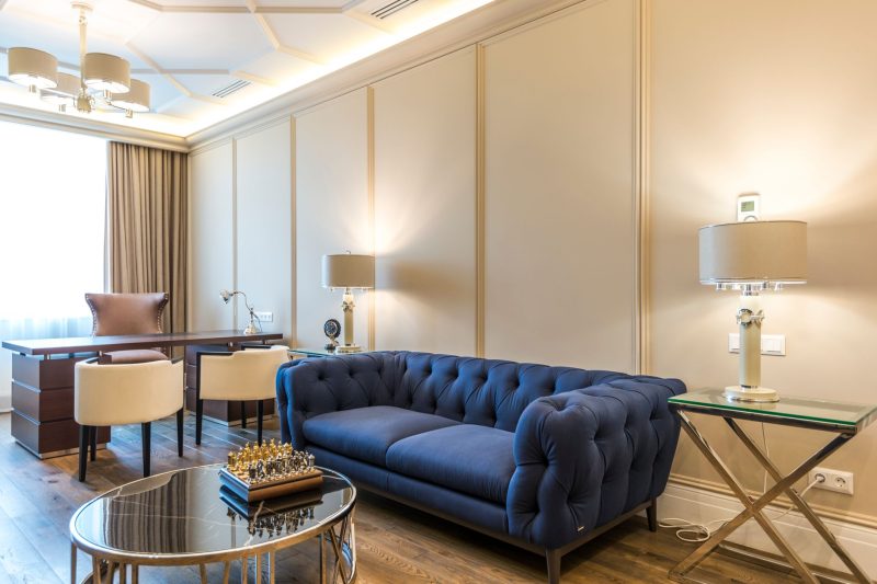 A modern living room with a blue tufted sofa, glass coffee table with a chess set, desk with white chairs, and two table lamps, all in a bright, well-lit space with beige walls and wood flooring.