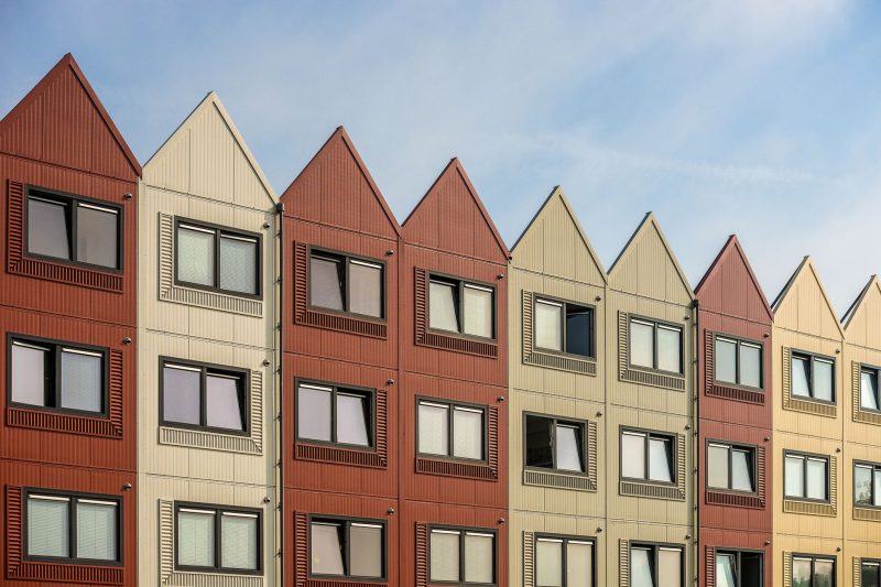 A row of modern apartment buildings with pointed roofs and alternating red and beige facades, featuring multiple rectangular windows under a partly cloudy sky.