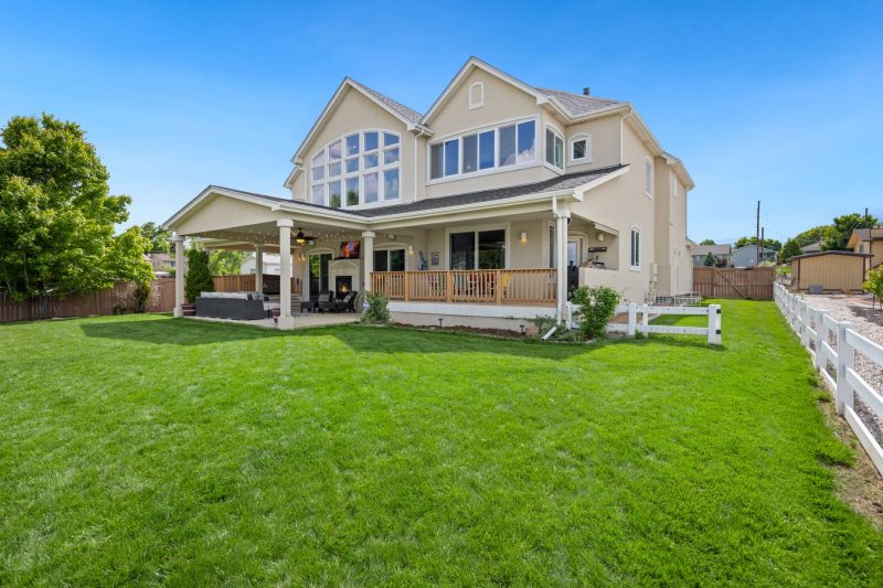 A two-story beige house with large windows, a covered patio, and a spacious green lawn, surrounded by a white fence and trees under a clear blue sky.