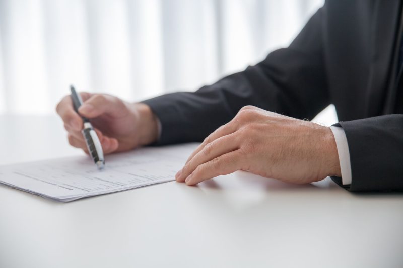 A person in a suit is holding a pen and filling out or signing a document on a white desk.