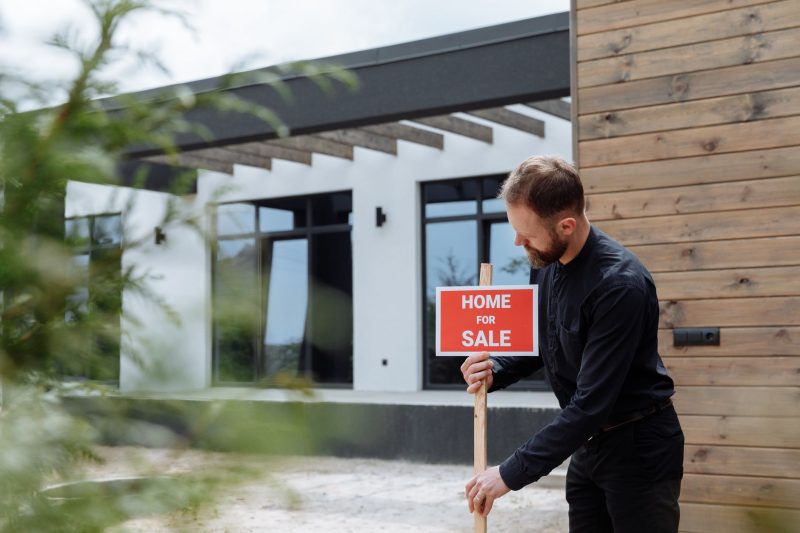 A man in a black shirt places a red Home For Sale sign in front of a modern house with large windows and wooden siding.