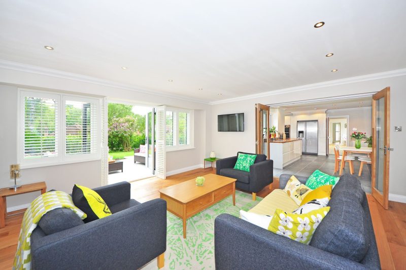 Bright living room with gray sofas, green and yellow pillows, a wooden coffee table, and open doors leading to a garden. Kitchen and dining area are visible in the background.
