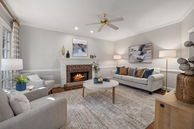 Modern living room with light gray sofas, a round coffee table, a brick fireplace with a fire burning, abstract art on the walls, large rug, and a ceiling fan. Natural light streams in from windows on the left.