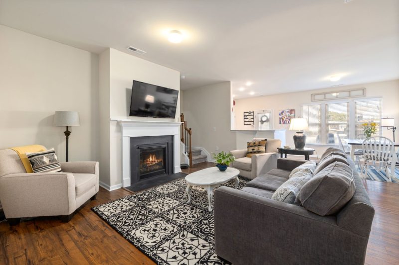 A cozy living room with a fireplace, TV, gray couch, armchair, patterned rug, and coffee table. The space opens to a dining area with a table, chairs, and large windows letting in natural light.