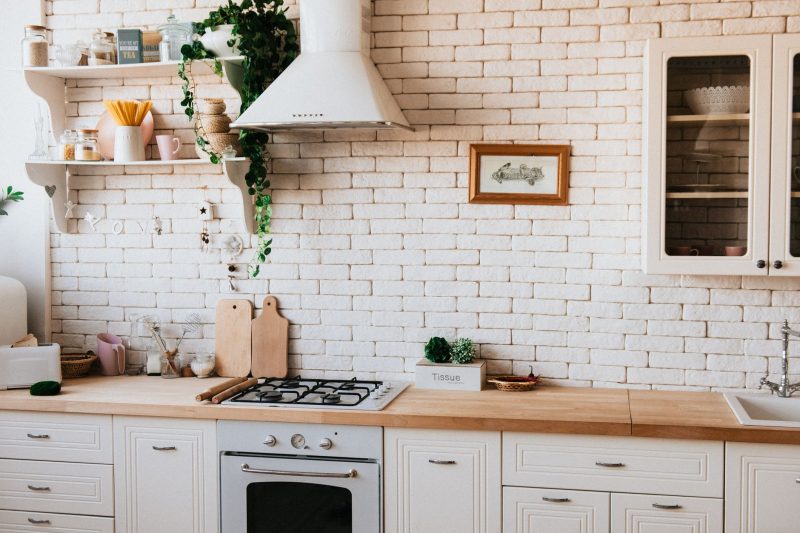 Bright kitchen with white brick walls, wooden countertops, white cabinets, a gas stove, shelves with plants and dishes, a range hood, and a framed picture on the wall.