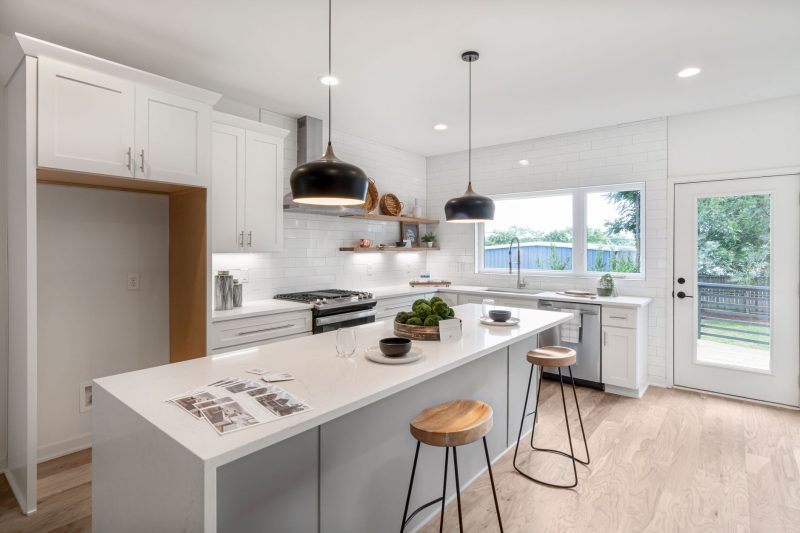A bright kitchen with white cabinets, a long quartz island with two bar stools, black pendant lights, and a white subway tile backsplash.
