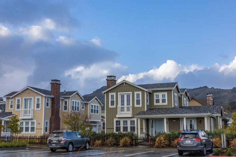 Two suburban houses with beige and olive siding, each with a car parked in front, under a cloudy sky.