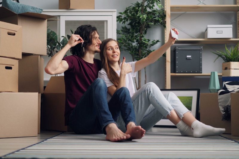 A couple sits on the floor surrounded by cardboard moving boxes as they take a selfie, with the man holding up a set of keys.